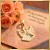 Złota zawieszka Aniołek Stróż obok herbacianych róż i napisu Kruchy jak płatek róży, ale chroniony przez anielskie skrzydła.jpg